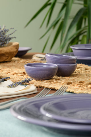 A close-up of a dining table features Fiesta’s Lavender 3 3/4 Inch Mini Bowl (5 OZ) and purple ceramic plates on a woven placemat, with silverware, napkins, lavender sprigs, and a green plant completing the inviting scene.
