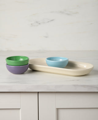 The Fiesta Lavender Bloom Mixed Color 4-Piece Condiment Set includes small green, purple, and blue bowls. On a white marble countertop, the green and purple bowls are stacked, and the blue bowl sits on an oval cream-colored serving tray.