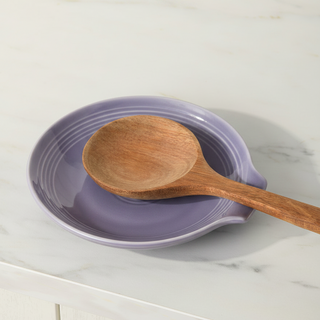 A wooden spoon rests on a Fiesta Lavender Extra Large Spoon Rest 7 Inch, a spacious purple utensil holder set on a white marble kitchen countertop.