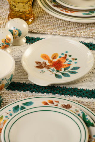 A decorative plate adorned with autumn leaves and acorns sits on a woven placemat, surrounded by coordinating dishes and fall-themed salt and pepper shakers. The look is completed with the Fiesta Autumn Fields Extra Large Spoon Rest 7 Inch, adding a charming and functional accent to your fall table decor.