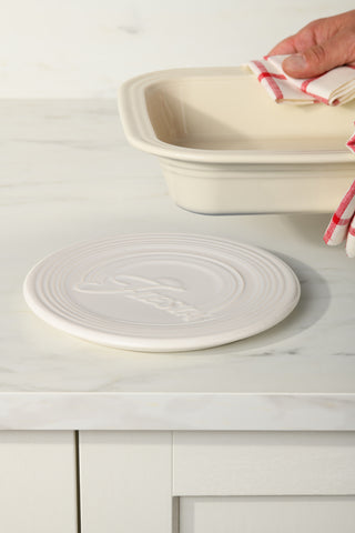 A person holds a ceramic baking dish on a white marble countertop with a red and white striped cloth, while an 8 1/4 Inch Extra Large Trivet from Fiesta adds both style and heat protection in the foreground.