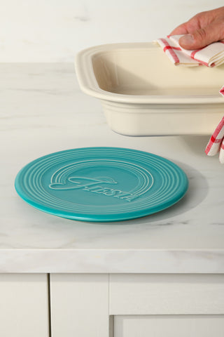 A hand lifts a beige baking dish near the Fiesta Extra Large Trivet 8 1/4 Inch, a heat-resistant ceramic trivet, on a white kitchen counter.