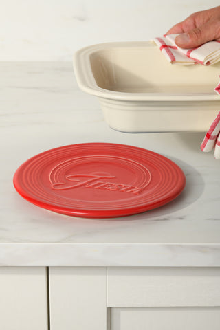 A Fiesta Extra Large Trivet 8 1/4 Inch in red, embossed with “Fiesta,” sits on a white countertop. Behind it, a hand with a red-striped towel lifts a cream baking dish. This ceramic trivet is stylish, sturdy, and heat-resistant.