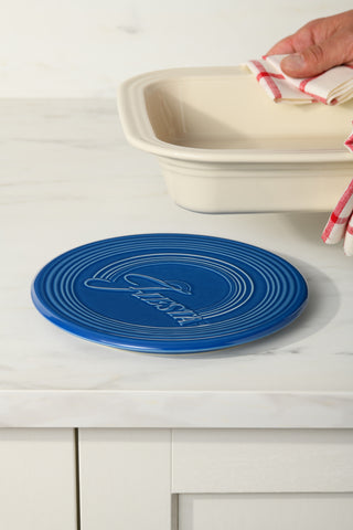A hand with a red-and-white striped towel holds a cream dish above the Fiesta Extra Large Trivet 8 1/4 Inch, a heat-resistant ceramic trivet embossed with "Fiesta," on a white marble countertop.