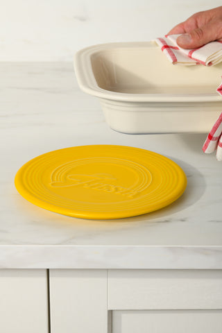 A hand holds a dish with a red and white towel, about to set it on the Fiesta Extra Large Trivet 8 1/4 Inch, which rests on a white marble countertop.