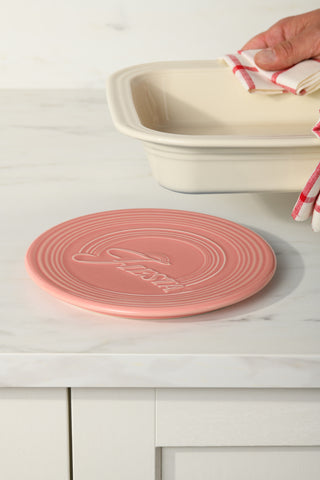 A hand holds a red and white striped towel near a cream baking dish on a marble counter, with the Fiesta Extra Large Trivet 8 1/4 Inch featured in the foreground.