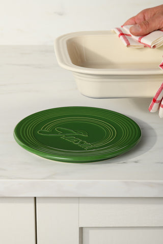 A hand with a red-striped towel lifts a cream-colored baking dish on a white countertop. In the foreground sits the Fiesta Extra Large Trivet 8 1/4 Inch, its textured design ideal for protecting surfaces from heat.