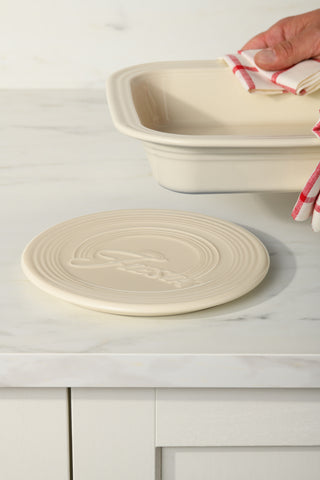 A cream-colored Fiesta Extra Large Trivet 8 1/4 Inch rests on a white marble countertop near a matching baking dish, while a hand holding the dish with a red and white striped towel stands out against the light cabinets in the background.