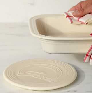 A hand holds a cream-colored Square Baker and Trivet with a red and white checkered cloth, positioned above the matching circular trivet on a marble surface.