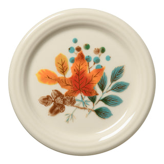 The Fiesta (FTC) Autumn Fields 4 3/8 Inch Coaster and Mug Cover is a round white ceramic piece featuring colorful autumn leaves, acorns, and berries on one side. Made in the USA from vitrified china; dishwasher safe for easy cleaning.
