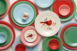 A vibrant assortment of plates and bowls in red, green, blue, and pink tones are artfully arranged, while some white plates showcase a charming red cardinal bird perched among branches with berries and leaves for a festive flair. Completing this cheerful holiday table setting is the Jolly Christmas Cardinal 4 3/8 Inch Coaster and Mug Cover by Fiesta from the Jolly Christmas Collection, adding an extra touch of seasonal joy.