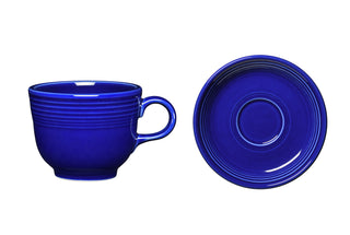 The Retired Twilight Cup and Saucer by Fiesta Factory Direct features a cobalt blue cup with a handle and matching saucer, both with subtle ridged detailing. The cup stands upright beside the saucer, shown from above in this elegant set.