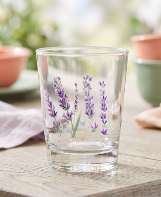 A set of four Moderne Glass Fiesta Lavender Bloom 15 oz. Tapered Double Old Fashion glasses, made in the USA and adorned with painted lavender flowers, sits on a wooden table with blurred Fiesta Dinnerware plates, bowls, and greenery in the background.