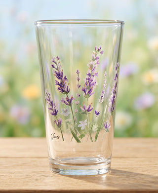 A set of four 16 oz. Moderne Glass Fiesta Lavender Bloom Cooler Glasses rests on a wooden surface, each piece adorned with painted lavender flowers and green stems. The softly blurred pastel background evokes a garden atmosphere. These charming glasses complement your Fiesta Dinnerware collection and are proudly made in the USA.