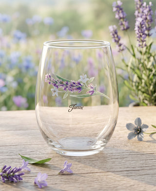 A set of four clear Fiesta Lavender Bloom 15 oz. Frame Stemless Glasses by Moderne Glass features a painted lavender sprig and the Fiesta logo, displayed on a wooden table surrounded by fresh lavender flowers with a garden softly blurred in the background.