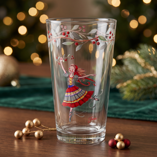 The Moderne Glass Nutcracker The Ballerina 16 oz. Cooler Glass - Set of 4 showcases a beautifully painted ballerina in a vibrant dress holding holly berries, surrounded by festive red berries and green leaves along the rim. Holiday ornaments and glowing lights softly blur in the background, adding to the seasonal charm. Made in the USA, this delightful set is a perfect companion to your Fiesta Dinnerware for a cheerful holiday display.