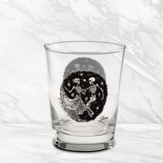 The Moderne Glass Fiesta Skeleton Duet 15 oz. Tapered Double Old Fashion glass features a black circular design with two dancing skeletons surrounded by small flowers. Made in the USA and inspired by Fiesta Dinnerware, this glass is pictured on a white marble surface.