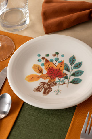 The Fiesta (FTC) Autumn Fields Classic Rim 7 1/4 Inch Salad Plate, featuring colorful autumn leaf and botanical designs, is displayed on an orange and green placemat with a fork, spoon, knife, glass, and orange napkin.