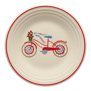 The Fiesta Americana Bicycle Classic Rim 9 Inch Luncheon Plate is a round, cream-colored vitrified china plate with a red rim. It features a red bicycle with blue and red streamers, a basket of red flowers, is dishwasher safe, and made in the USA.
