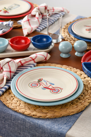 A close-up of a dining table set with woven placemats, Fiesta Americana Bicycle Classic Rim 9 Inch Luncheon Plates, colorful bowls, checkered napkins, glasses, and light blue salt and pepper shakers for a charming touch.