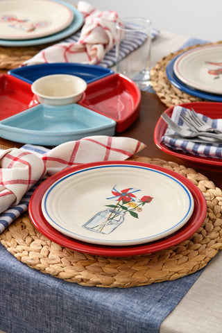 A table set for a meal features woven placemats, Fiesta Americana Mason Jar Classic Rim 9-Inch Luncheon Plates, striped and plaid cloth napkins, and blue and red divided serving trays—all thoughtfully made in the USA.