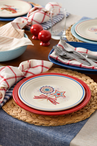 A festive table features a woven placemat, Fiesta Americana Fan, Star, & Bow Classic Rim 9 Inch Luncheon Plate with patriotic designs, red and white napkins, cutlery, glasses, and red salt and pepper shakers on a wooden table.
