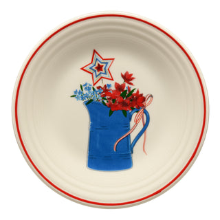 The Fiesta Americana Farmhouse Pitcher Classic Rim 9 Inch Luncheon Plate features a cream base, red edge, and a blue pitcher with red and blue flowers, star accent, and a red-and-white striped ribbon on the handle.