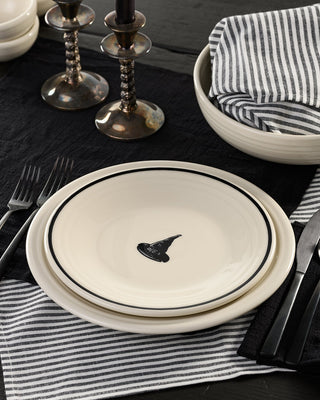 The Fiesta Halloween Icon Witch’s Hat Classic Rim 9 Inch Luncheon Plate, featuring a striking black design on crisp white, sits elegantly atop a black placemat surrounded by striped napkins, silverware, and two candlesticks—perfect for adding a festive touch to your Halloween table setting.