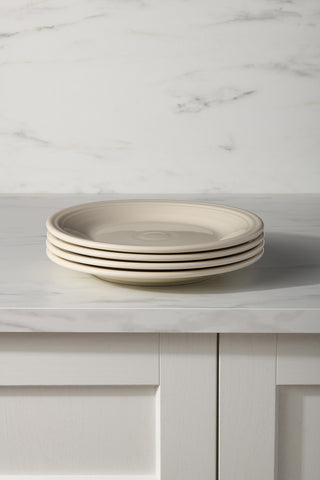 A set of four Fiesta (FTC) Linen Classic Rim 10 1/2 Inch Dinner Plates are stacked neatly on a white marble countertop, with sleek white cabinets and a marble-patterned wall in the background.