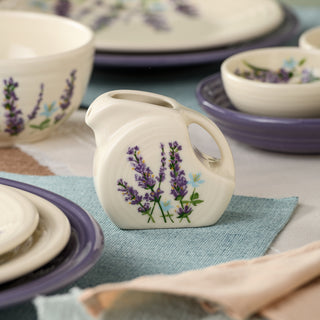 The Fiesta Lavender Bloom 5 OZ Mini Disk Pitcher, adorned with painted lavender flowers and crafted from lead-safe china, features a convenient handle and rests on a set table alongside matching floral dishes and pastel linens—making it a perfect collectible serveware piece.