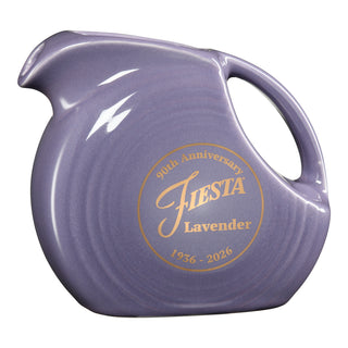The Fiesta 2026 Lavender Anniversary Small Disk Pitcher by Fiesta is a USA-made, lavender pitcher with a round body, wide handle, and gold "90th Anniversary FIESTA Lavender 1936-2026" text on its side.