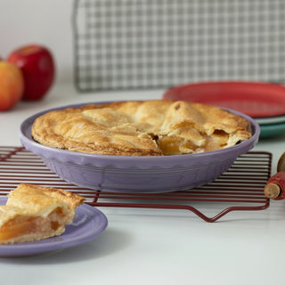 A golden-brown apple pie rests in the Fiesta Lavender 10 1/4 Inch Deep Dish Pie Baker (44oz), an oven-safe purple dish on a cooling rack, with a slice on a matching plate; stacked plates and apples appear in the background.