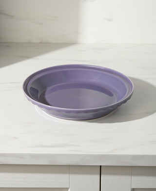 A round, empty Fiesta Lavender 10 1/4 Inch Deep Dish Pie Baker (44oz) rests on a white marble countertop in a bright kitchen. This oven safe ceramic dish is made in the USA.