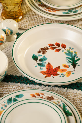 A close-up of the Fiesta Autumn Fields 10 1/4 Inch Deep Dish Pie Baker (44oz), accented with autumn leaves and acorns, is surrounded by coordinating fall tableware and a salt and pepper shaker on a woven tablecloth, showcasing the collection’s vibrant seasonal colors.