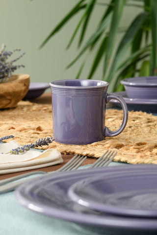 A Fiesta Lavender 12 OZ Java Coffee Mug sits on a woven placemat with matching plates, utensils, and a cloth napkin, accented by lavender sprigs and green plants in the background. Made in the USA.