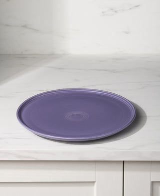The Fiesta Lavender 12 Inch Round Baking and Serving Platter rests on a white marble countertop with a subtle ring pattern, set against a white marble backsplash and white cabinetry.