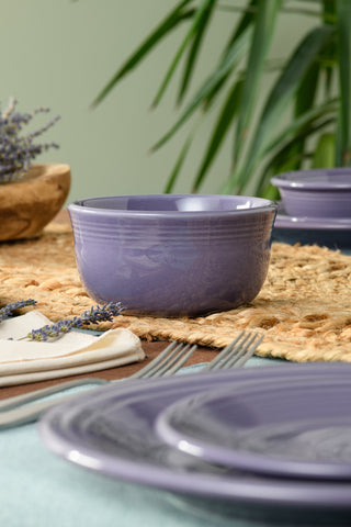 A Fiesta Lavender All-Purpose 6 Inch Gusto Bowl, 28 oz capacity, sits on a woven placemat among matching plates and silverware, accented by a sprig of lavender and green leaves in the background.