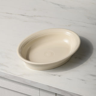 The Fiesta New Retired Linen Oval Baker, 10 1/4 inch and 40 oz, in cream ceramic sits on a white marble countertop. This oven-safe dish features subtle rim ridges and a concentric circle pattern at the bottom.