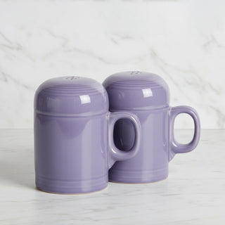 Two Fiesta Lavender 4 5/8 Inch Rangetop Large Salt and Pepper Shakers, made in the USA, sit upside down with outward-facing handles on a white marble surface. A white marble wall serves as the background.