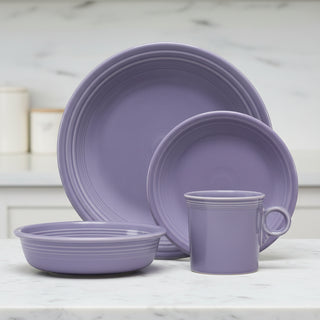 The Fiesta Lavender Classic Rim 4-Piece Dinnerware Set, Service for 1, features a dinner plate, salad plate, bowl, and vintage ring handle mug in a soft purple hue, beautifully arranged on a white countertop in a kitchen with a marble backsplash.