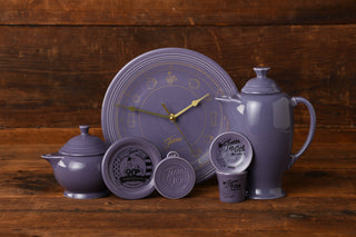 The Lavender 90th Anniversary Bundle by Fiesta Factory Direct, featuring beautifully crafted ceramic pieces—including a teapot, sugar bowl, cup, small containers, and a clock-shaped plate handmade in the USA—is displayed on a wooden surface.
