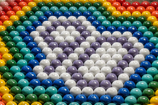 Rows of colorful plastic balls with small holes are arranged in concentric circles, forming a pattern with green, yellow, red, blue, purple, and white sections. The overall design creates a visually striking, symmetrical display.