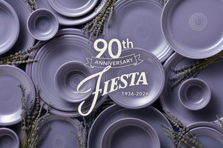 A flat-lay arrangement of lavender-colored ceramic plates and bowls with a central white script overlay reading '90th Anniversary Fiesta 1936-2026,' decorated with sprigs of lavender.