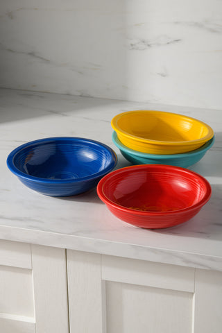 A set of four Fiesta Mixed Vibrant Classic Rim 8 3/8-Inch 34OZ Pasta Bowls—blue, red, yellow, and green—are stacked on a white marble countertop. Dishwasher safe, these bowls bring lively color to your kitchen.