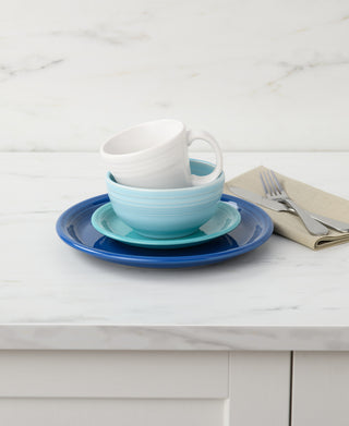 A stack of blue and white ceramic dishes, including a plate, bowl, saucer, and a Fiesta 15 OZ Tapered Mug by Fiesta made in the USA, sits on a white marble countertop beside a beige napkin with a fork and knife on top.