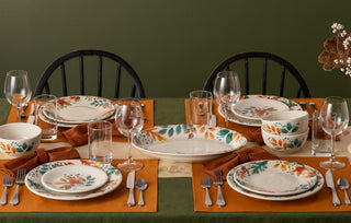A dining table set for four with autumn-themed plates, orange napkins, drinking glasses, and silverware on orange placemats. The table has a green tablecloth and two wooden chairs are visible against a green wall.