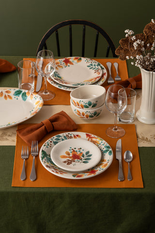 A dining table for two with Fiesta’s Autumn Fields Classic Rim 10 1/2 Inch Dinner Plates, orange napkins, silver cutlery, clear glasses, a white floral vase, and a green-orange tablecloth creates a cozy fall atmosphere.