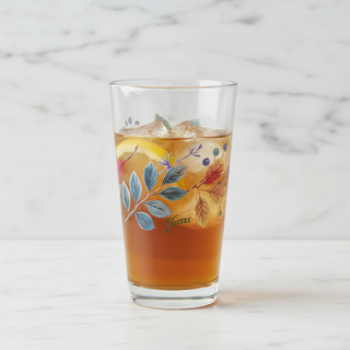 A clear pint glass with an autumn leaf and berry illustration, filled with an iced amber beverage and a lemon slice, set on a white marble surface against a white marble background.