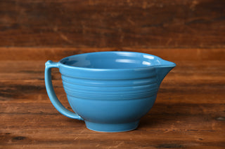 A large blue ceramic mixing bowl—the Fiesta Factory Direct Peacock Batter Bowl in the retired Peacock color—with a handle and spout sits on a wooden surface against a softly blurred wood background.