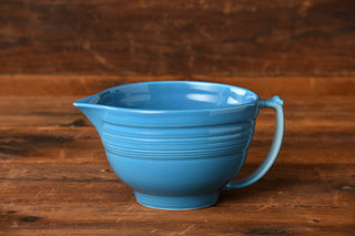 A retired Fiesta Factory Direct Peacock Batter Bowl with a handle and spout sits gracefully on a rustic wooden surface, set against a rich brown wooden background.